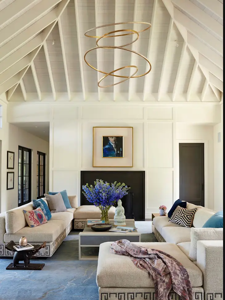 A modern living room with vaulted white ceiling, two beige sofas, blue accents, a central coffee table with flowers, abstract art, and a gold spiral chandelier.