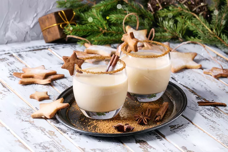 Two glasses of eggnog with cinnamon sticks and star-shaped cookies on a tray, surrounded by more cookies and holiday greenery.