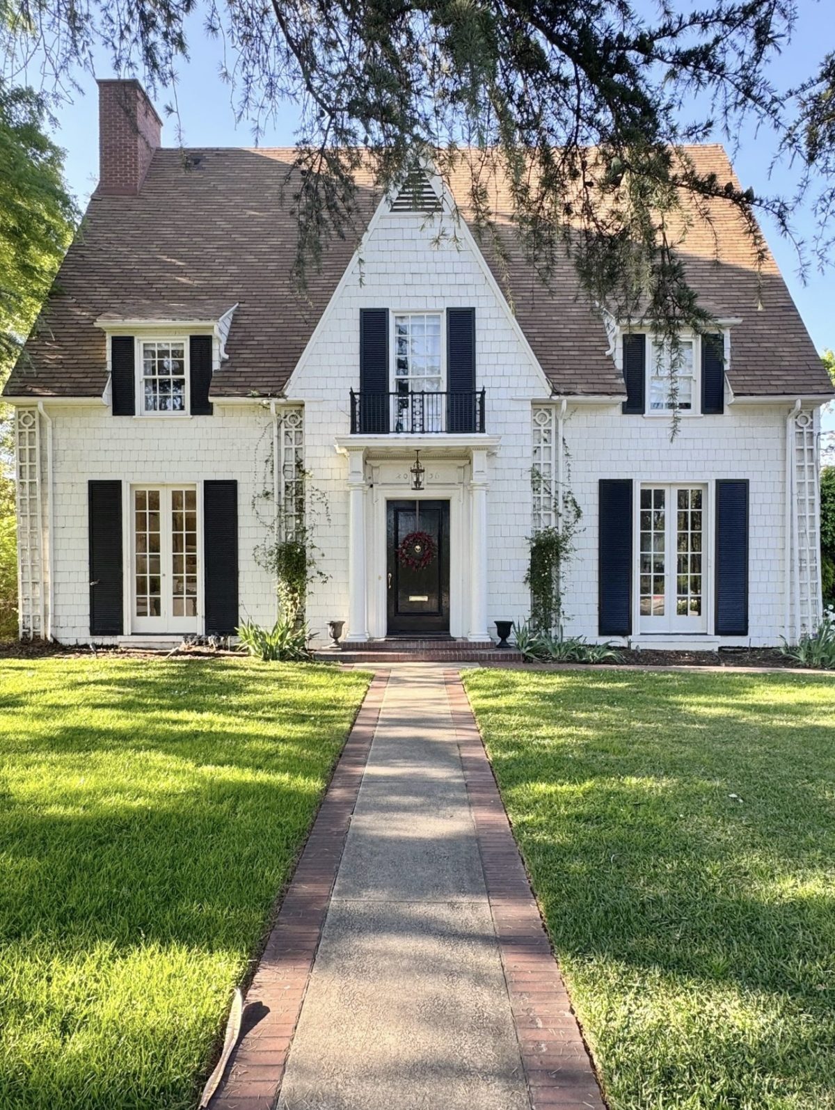 Two-story white house with steep roof, black shutters, central front door, small balcony, and a brick walkway leading across a manicured lawn.