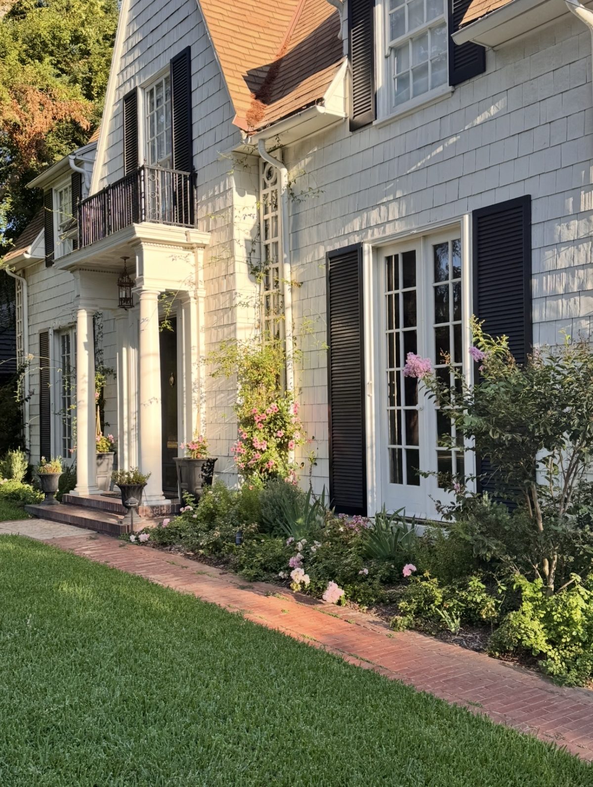 A two-story house with gray siding, black shutters, white trim, and a small front porch sits behind a brick walkway and a garden with green plants and blooming flowers.