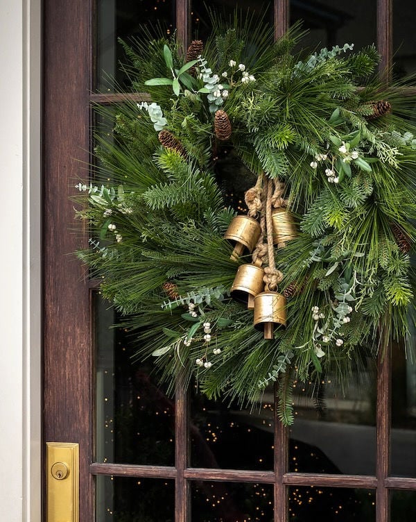A green holiday wreath with pine needles, pinecones, and small white berries hangs on a glass-paneled door, decorated with three brass bells tied with rope.
