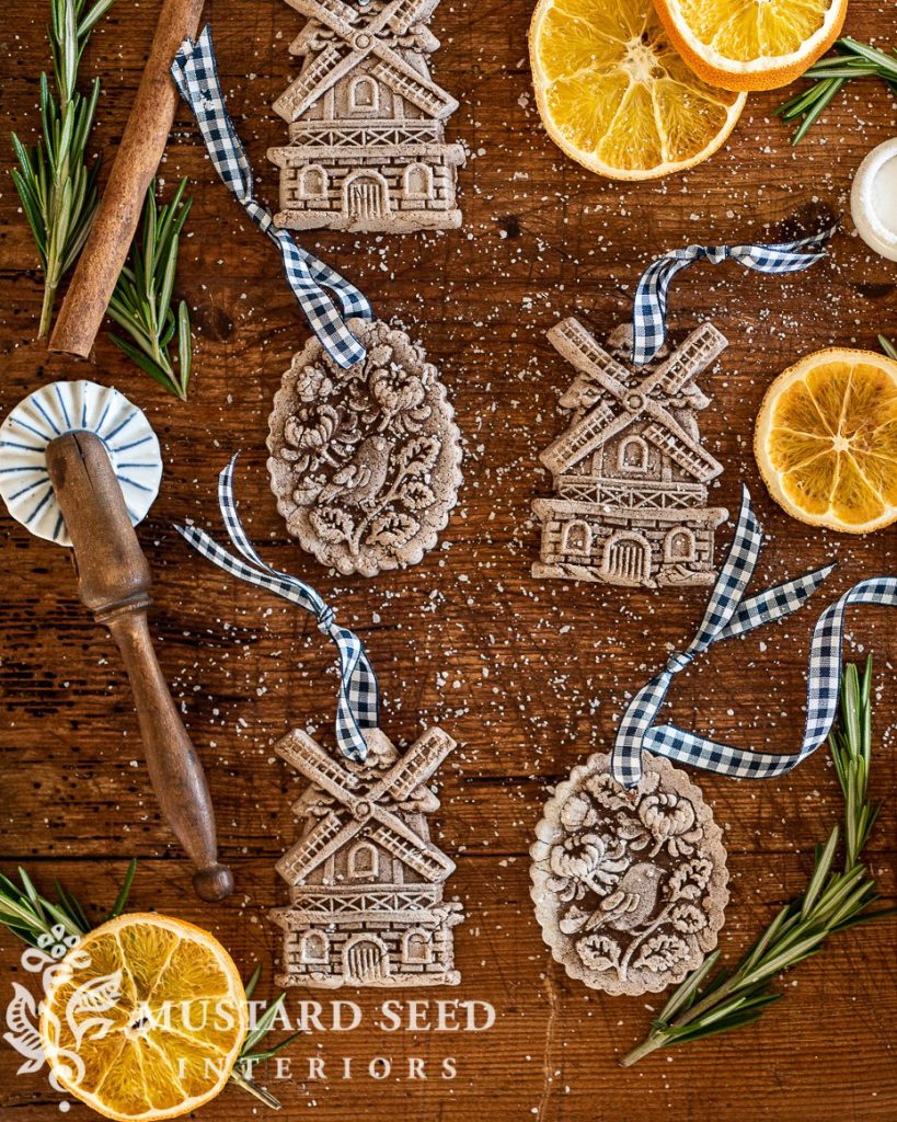Five intricately designed cookies with blue plaid ribbons, dried orange slices, rosemary sprigs, and baking tools on a wooden surface. "Mustard Seed Interiors" logo in the corner.