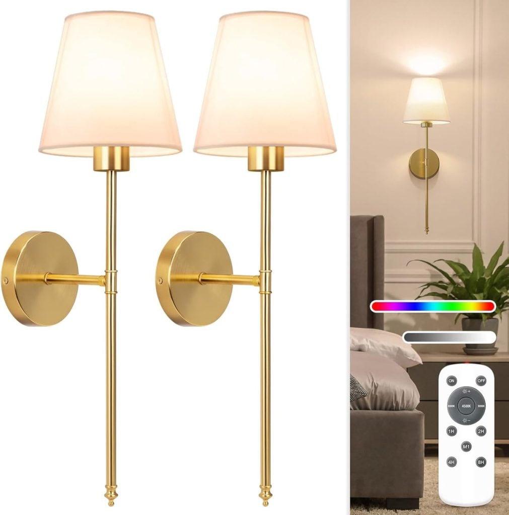 A pair of gold wall sconces with white shades, shown mounted above a bedside table with a plant. An Amazon Find, these sconces include a remote control for adjusting light color and brightness.