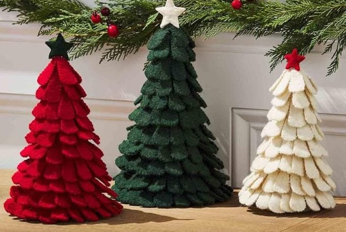 Three felt Christmas tree decorations in red, green, and white are displayed on a wooden table, with a pine garland and red berries on the railing behind them.
