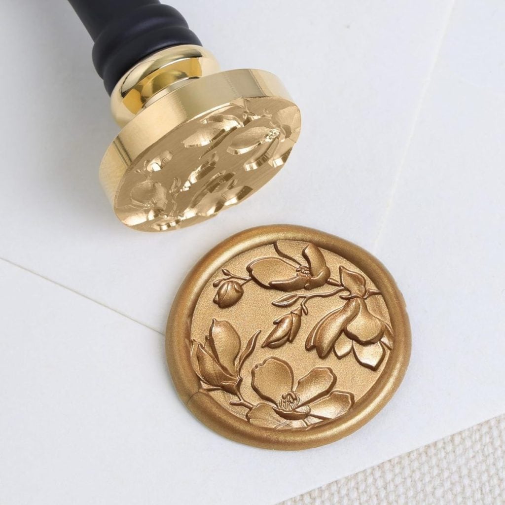 A gold wax seal with a floral design is pressed onto white paper using a brass stamp with a black handle.