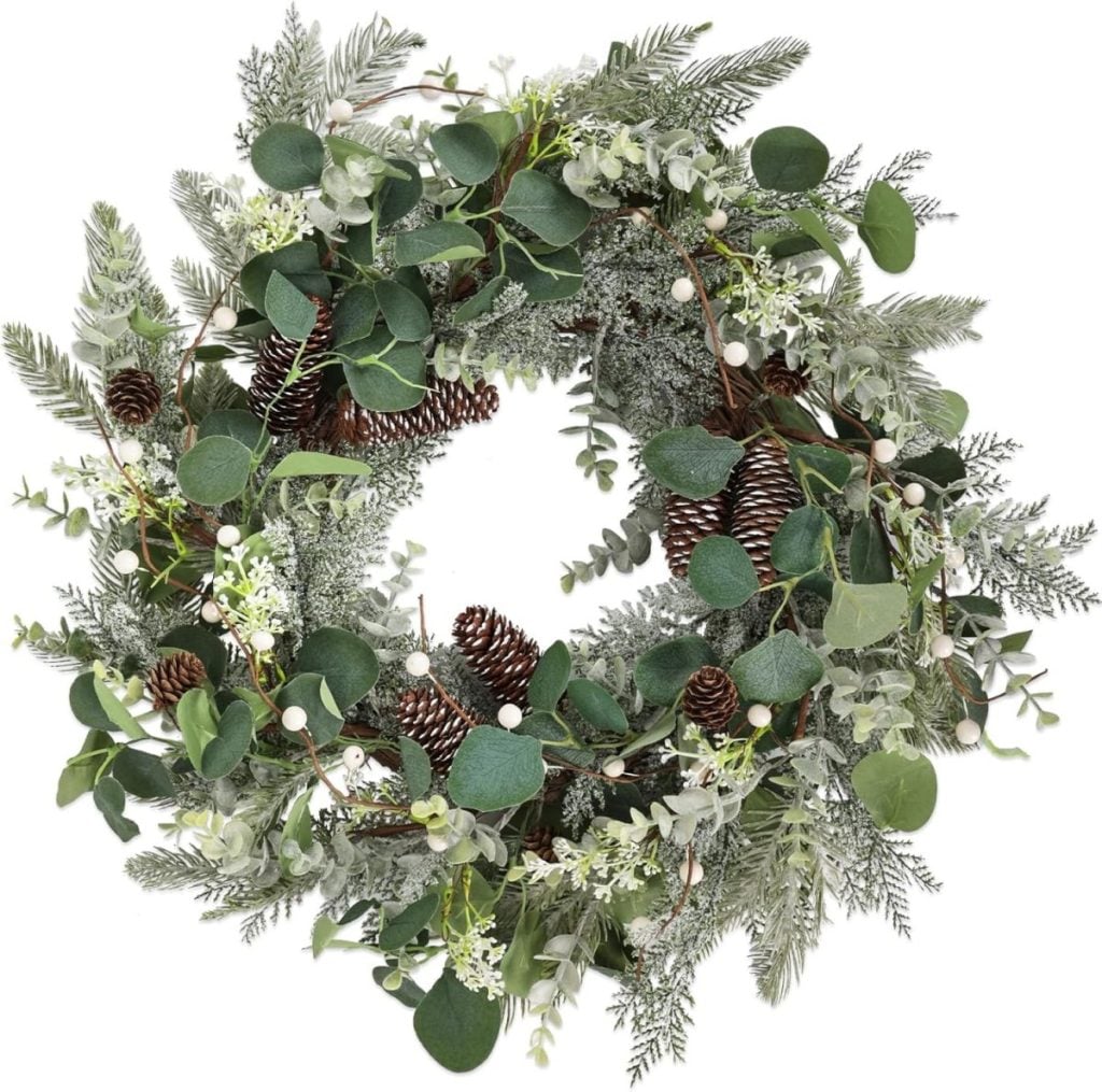 A decorative wreath made of green leaves, pine branches, small white berries, and pine cones arranged in a circular shape.
