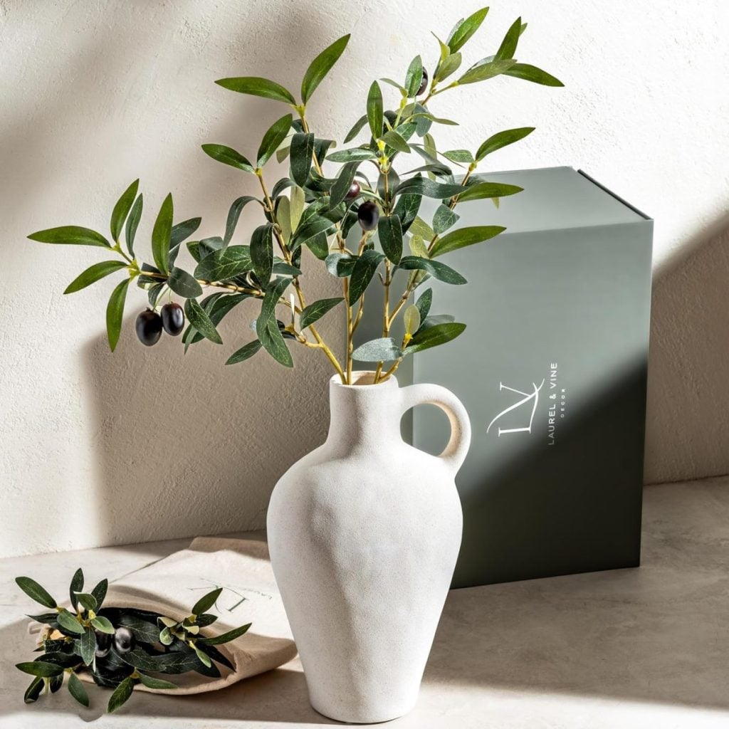 A white ceramic vase with olive branches sits on a light surface next to a green gift box and a small branch with olives and leaves.