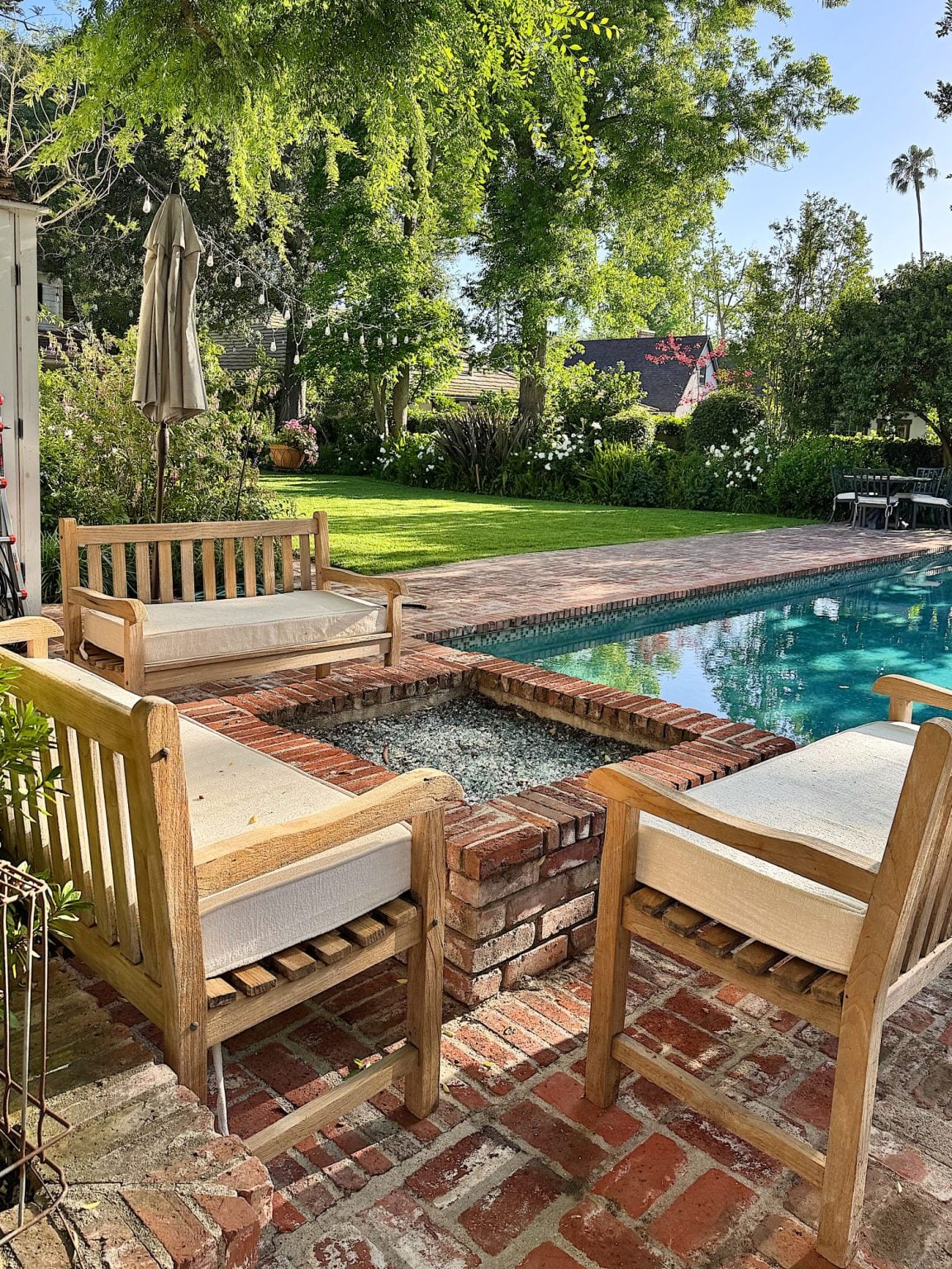 Two wooden benches and a chair surround a brick fire pit beside a swimming pool in a backyard with lawn and trees, featuring cozy Amazon Finds perfect for outdoor gatherings.