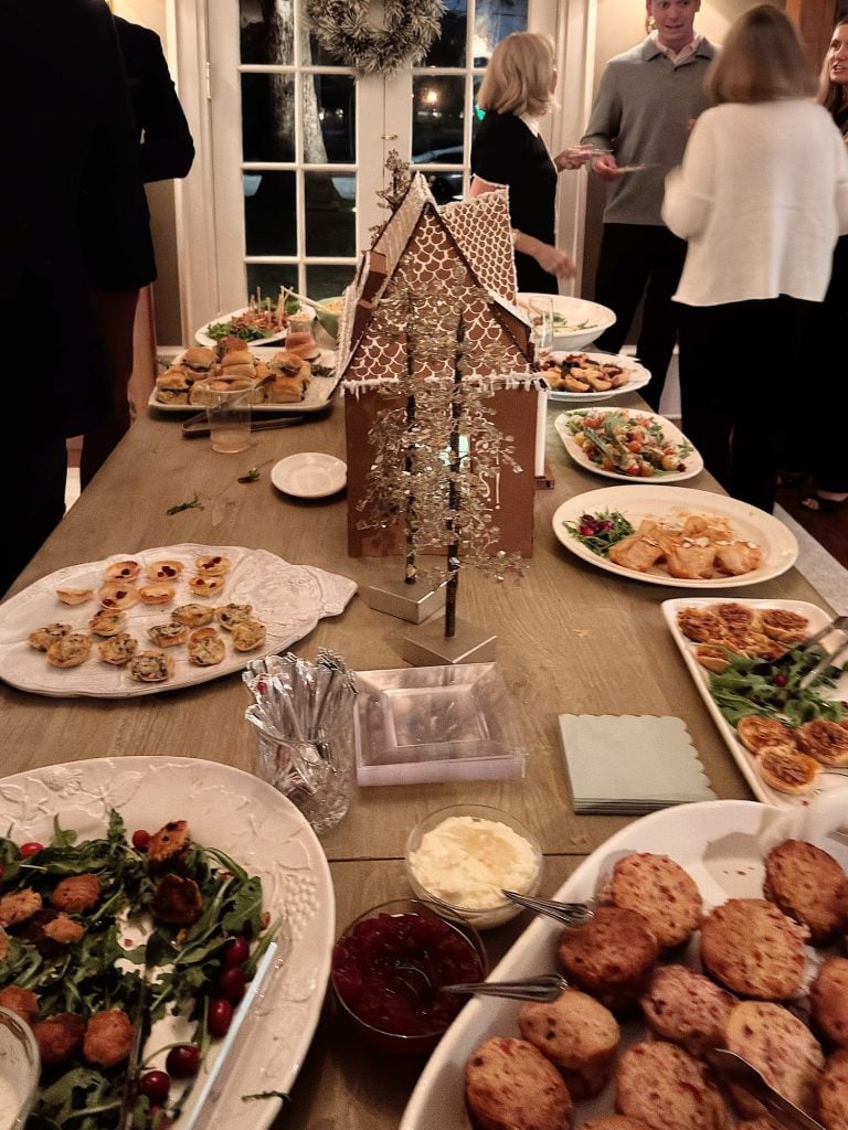 A buffet table with assorted appetizers and a gingerbread house centerpiece; people are gathered and serving themselves in the background.
