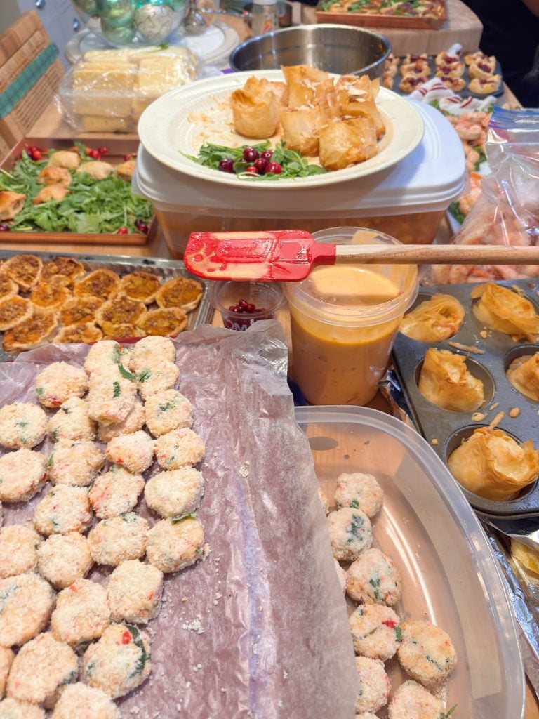 A variety of prepared and partially prepared appetizers on a busy kitchen counter, including breaded bites, pastry cups, and a container of dipping sauce with a spatula.