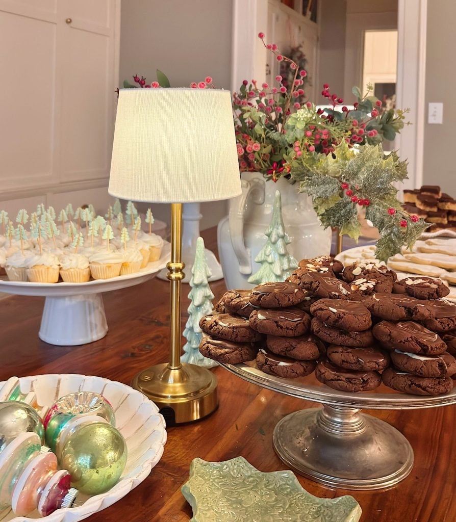 A table with chocolate cookies, cupcakes, layered bars, a lamp, ceramic Christmas trees, ornaments, and a floral arrangement set for a holiday gathering.