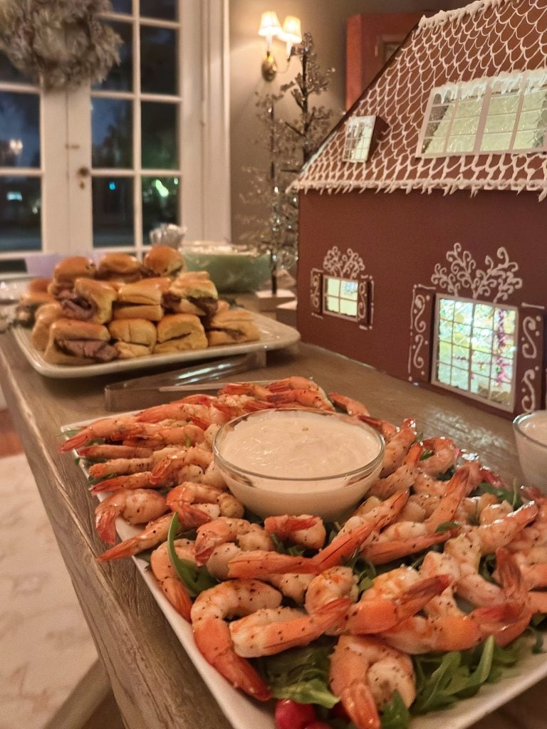 A platter of shrimp with dipping sauce sits on a table next to a plate of sandwiches and a large gingerbread house.