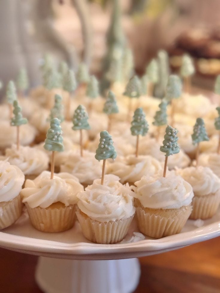 A plate of vanilla cupcakes with white frosting, each topped with a toothpick holding a small, blue-green decorative tree.