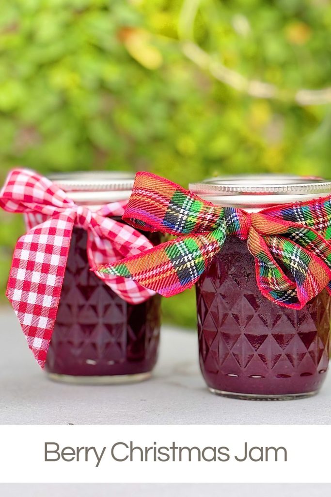 Two jars of berry jam with decorative holiday bows on the lids sit on a light surface, with a blurred green background. Text reads "Berry Christmas Jam.