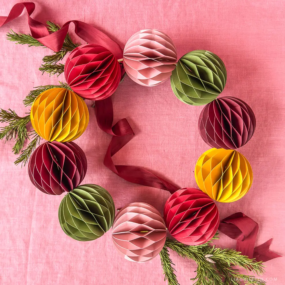 A wreath made of colorful honeycomb paper balls and greenery, arranged on a pink fabric background with a red ribbon.
