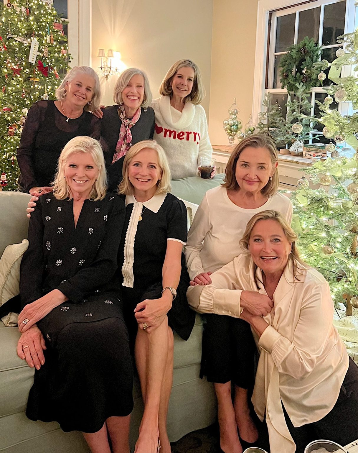 Seven women smile and pose together in a living room decorated with Christmas trees and holiday lights.