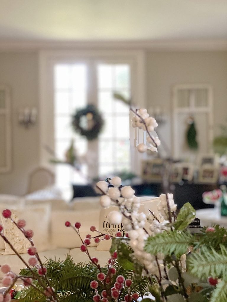 A blurred living room with holiday decorations, featuring berry branches and greenery in the foreground and a wreath on French doors in the background.