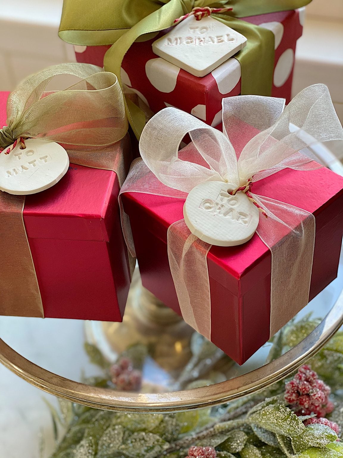 Three wrapped gift boxes with bows and name tags reading "Michael," "Matt," and "Char" are displayed on a glass surface with holiday greenery underneath.