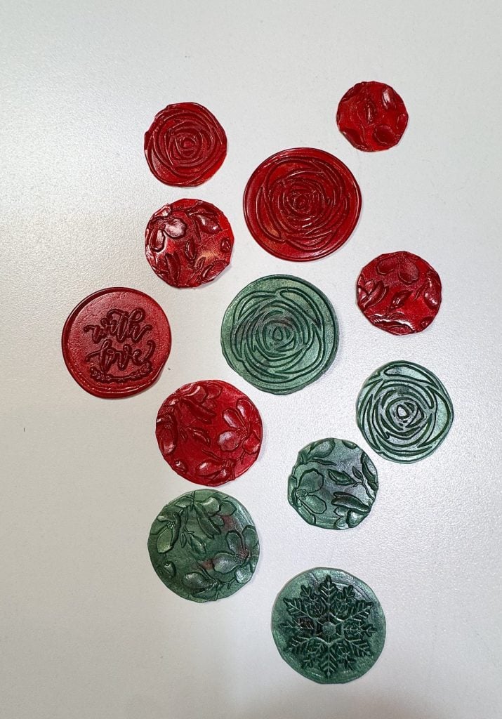 Thirteen red and green wax seals with various floral and leaf patterns are displayed on a white surface.