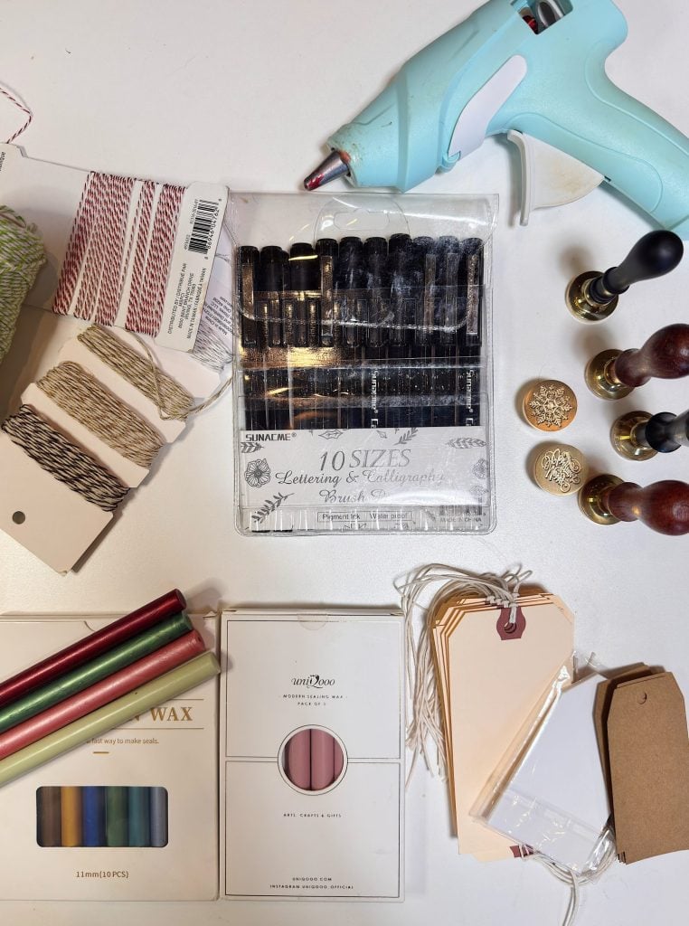A flat lay of crafting supplies including a blue glue gun, wax seals, calligraphy pens, tags, twine, wax sticks, and metallic rods arranged on a white surface.