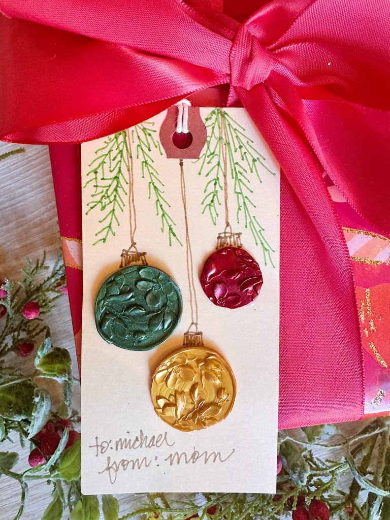 A gift wrapped in bright pink paper with a pink ribbon, featuring a tag decorated with green, red, and gold wax ornament seals, labeled "to: michael, from: mom.