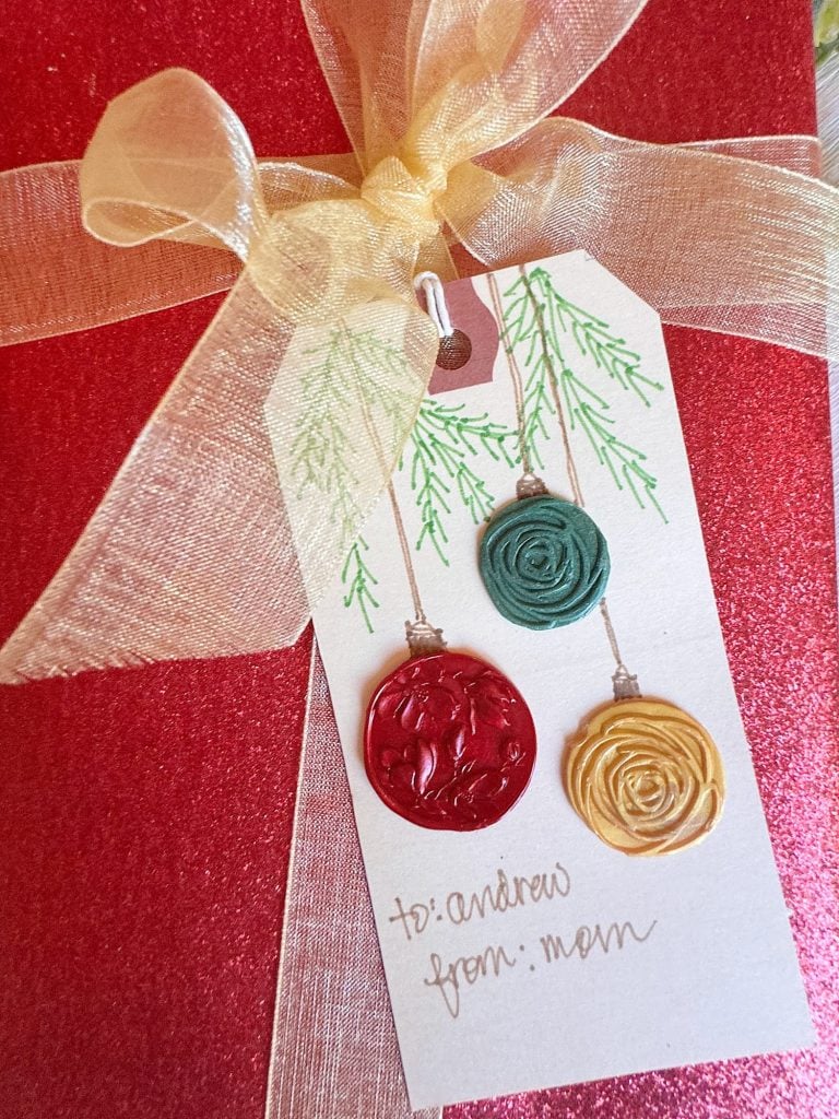 A red glitter gift box with a gold ribbon and a gift tag decorated with green branches, wax seals, and handwritten "to: andrew from: mom.