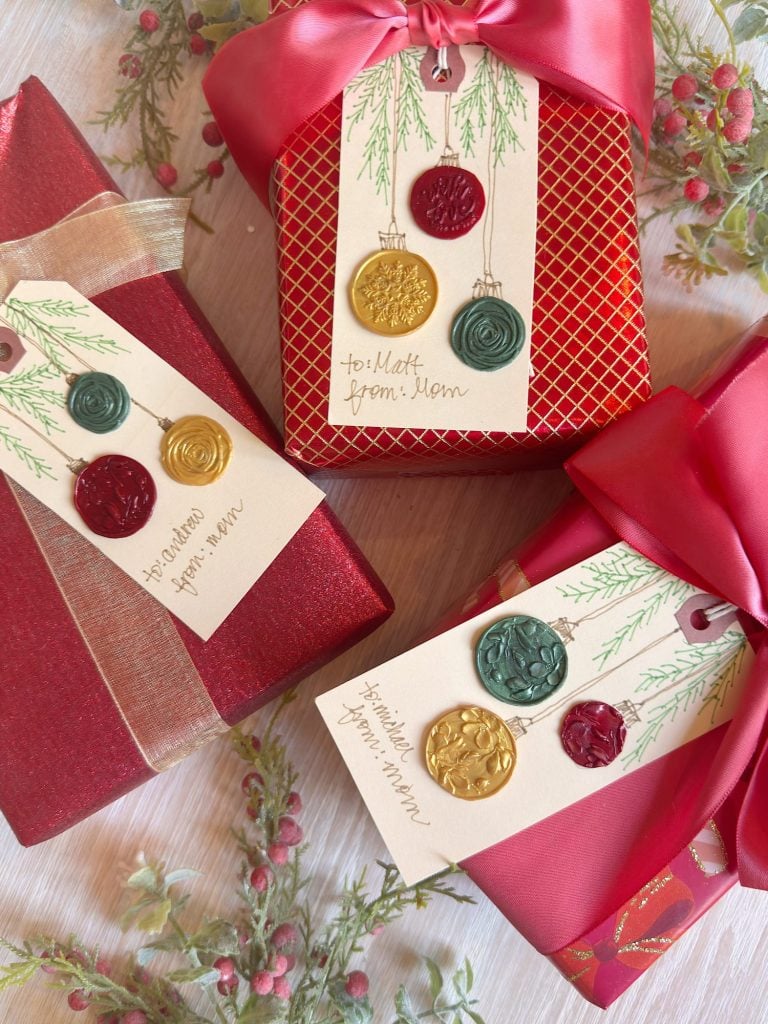 Three red gift boxes with festive ribbon and wax seal gift tags are arranged on a table decorated with holiday greenery and berries.
