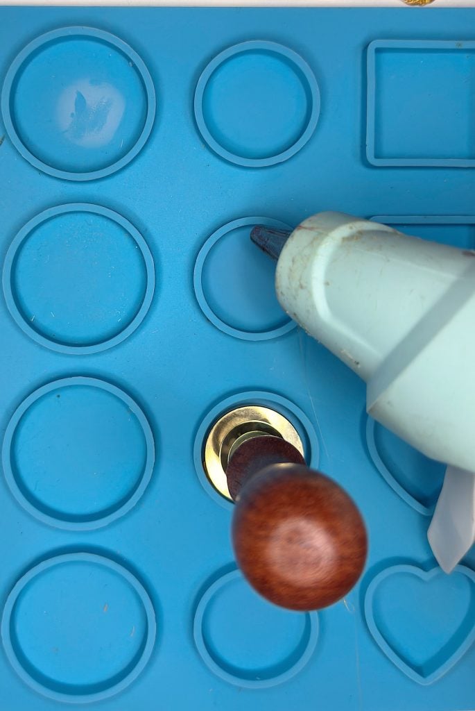 A hot glue gun and a wax seal stamp rest on a blue silicone mold with circular, square, and heart-shaped cavities.