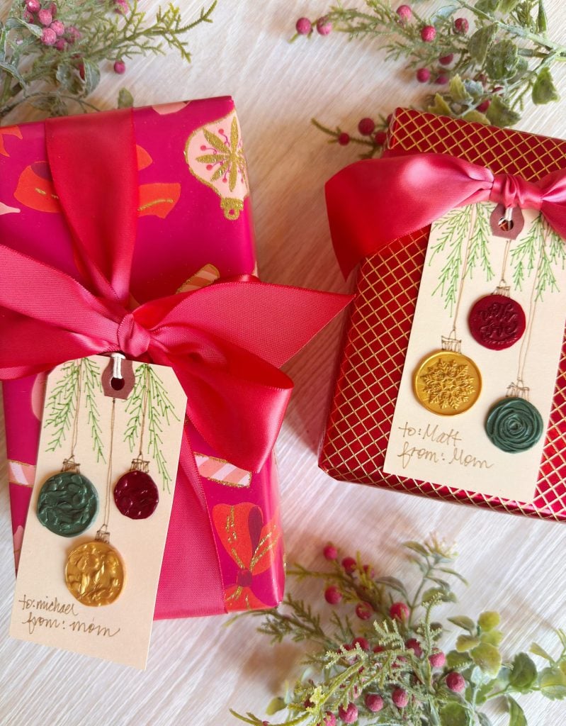 Two gift boxes with red ribbon and festive tags decorated with wax seals; the tags are labeled “to: Michael from: Mom” and “to: Matt from: Mom.” Berries and greenery surround the boxes.