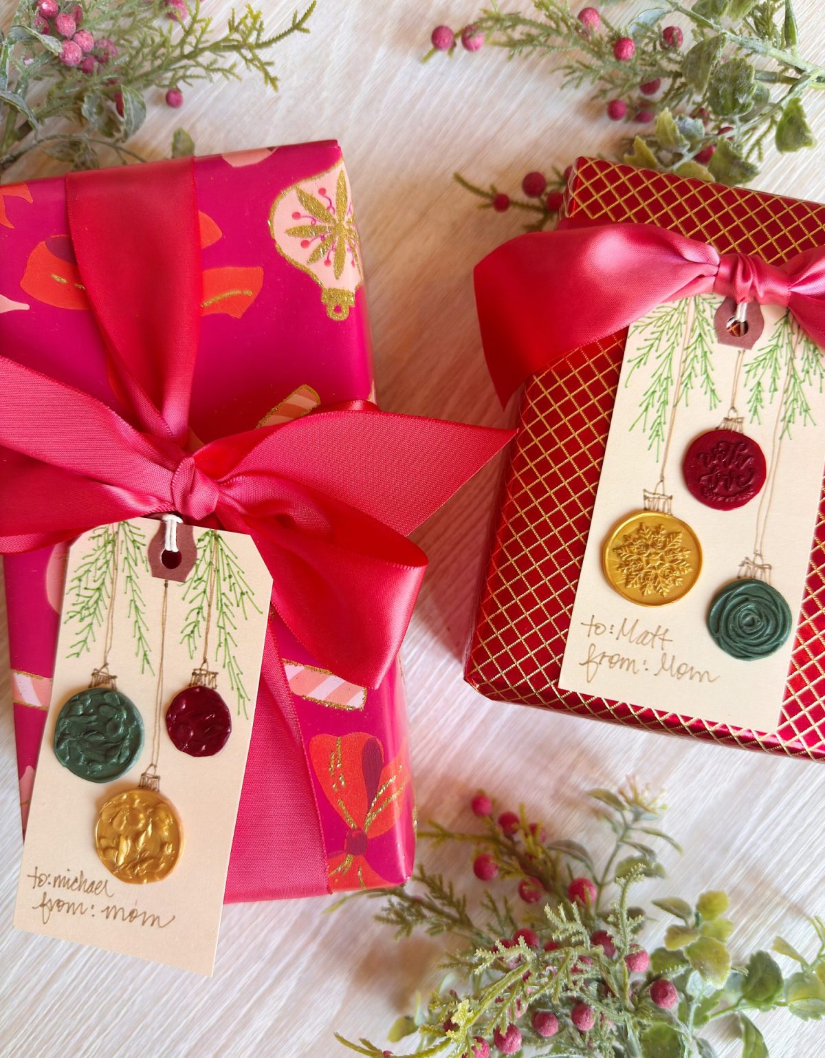 Two gift boxes with red ribbon and festive tags decorated with wax seals; the tags are labeled &ldquo;to: Michael from: Mom&rdquo; and &ldquo;to: Matt from: Mom.&rdquo; Berries and greenery surround the boxes.
