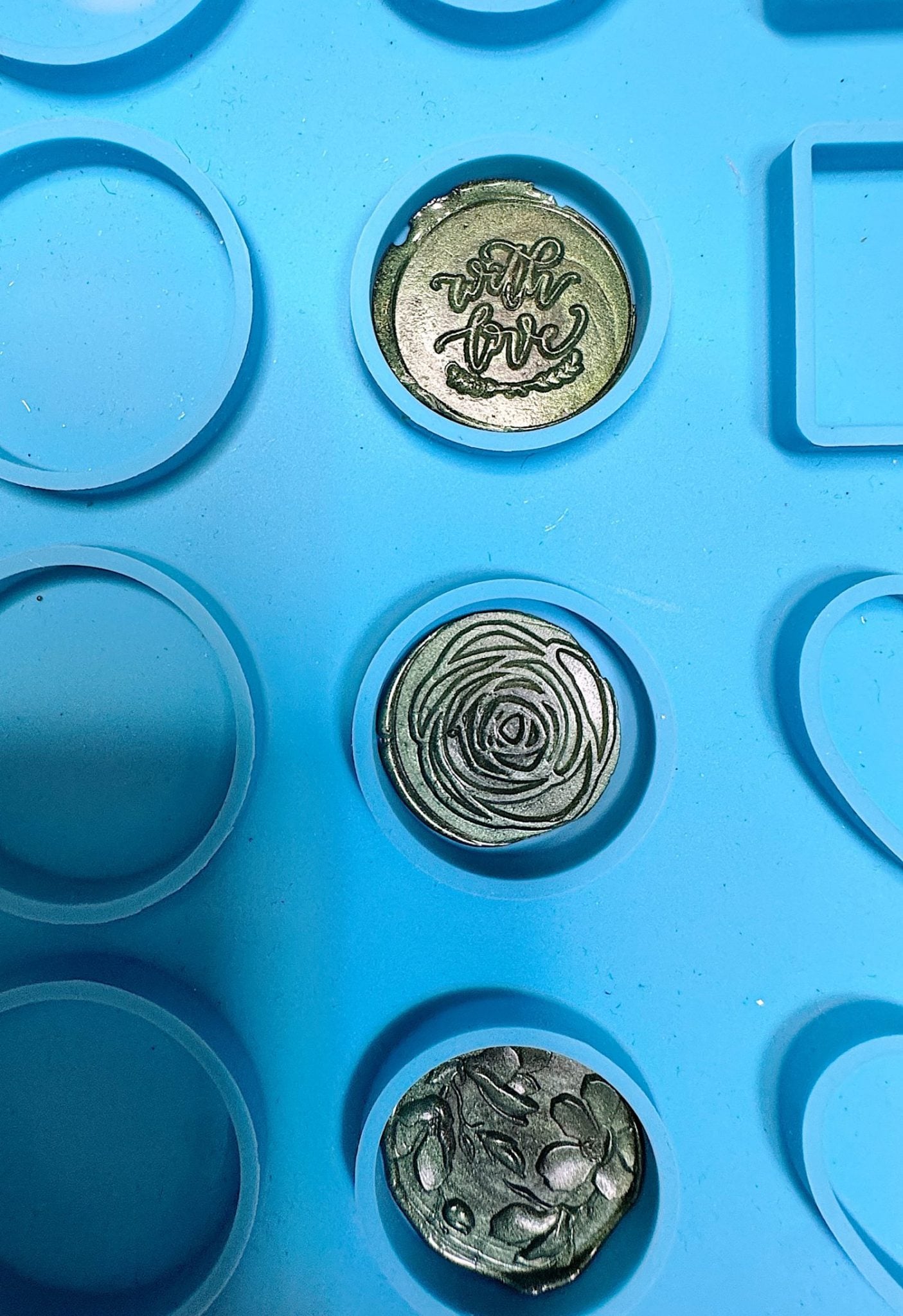 Three round metal stamps with different designs&mdash;"with love" text, a rose, and floral pattern&mdash;placed in a blue silicone mold with circular compartments.