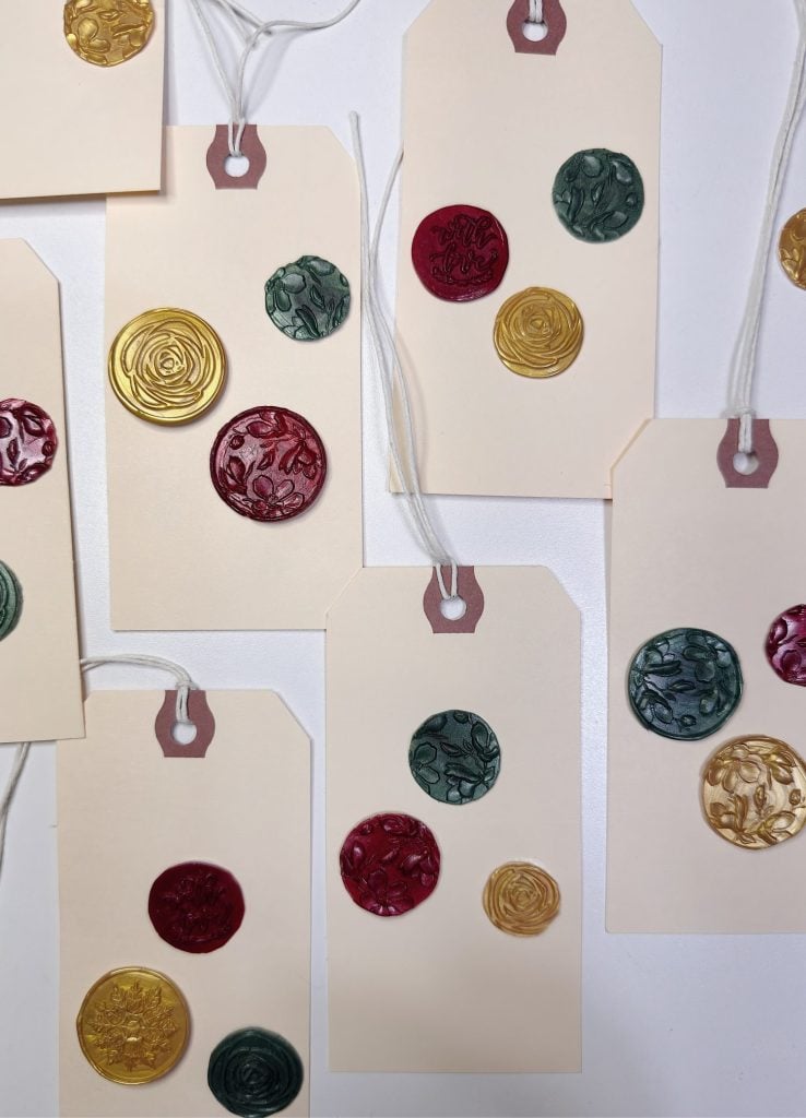 Gift tags with red, green, and gold wax seals are arranged on a white surface. Each tag has one or two wax seals and a white string attached.