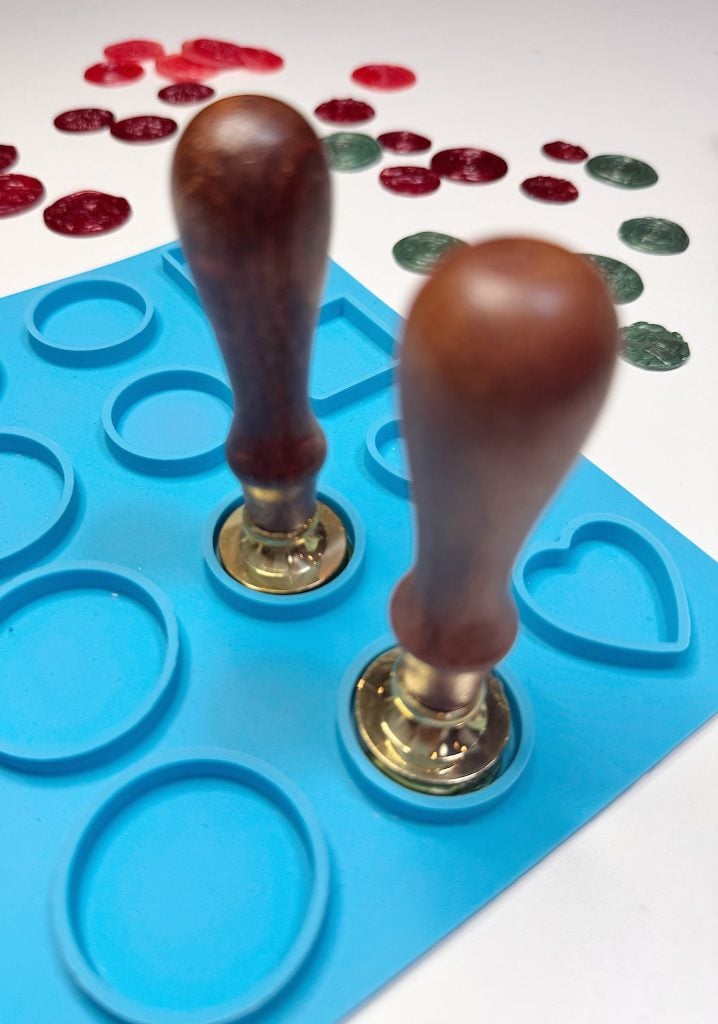 Two wooden-handled wax seal stamps rest on a blue silicone mold with circular and heart-shaped cavities, with wax seals scattered in the background.