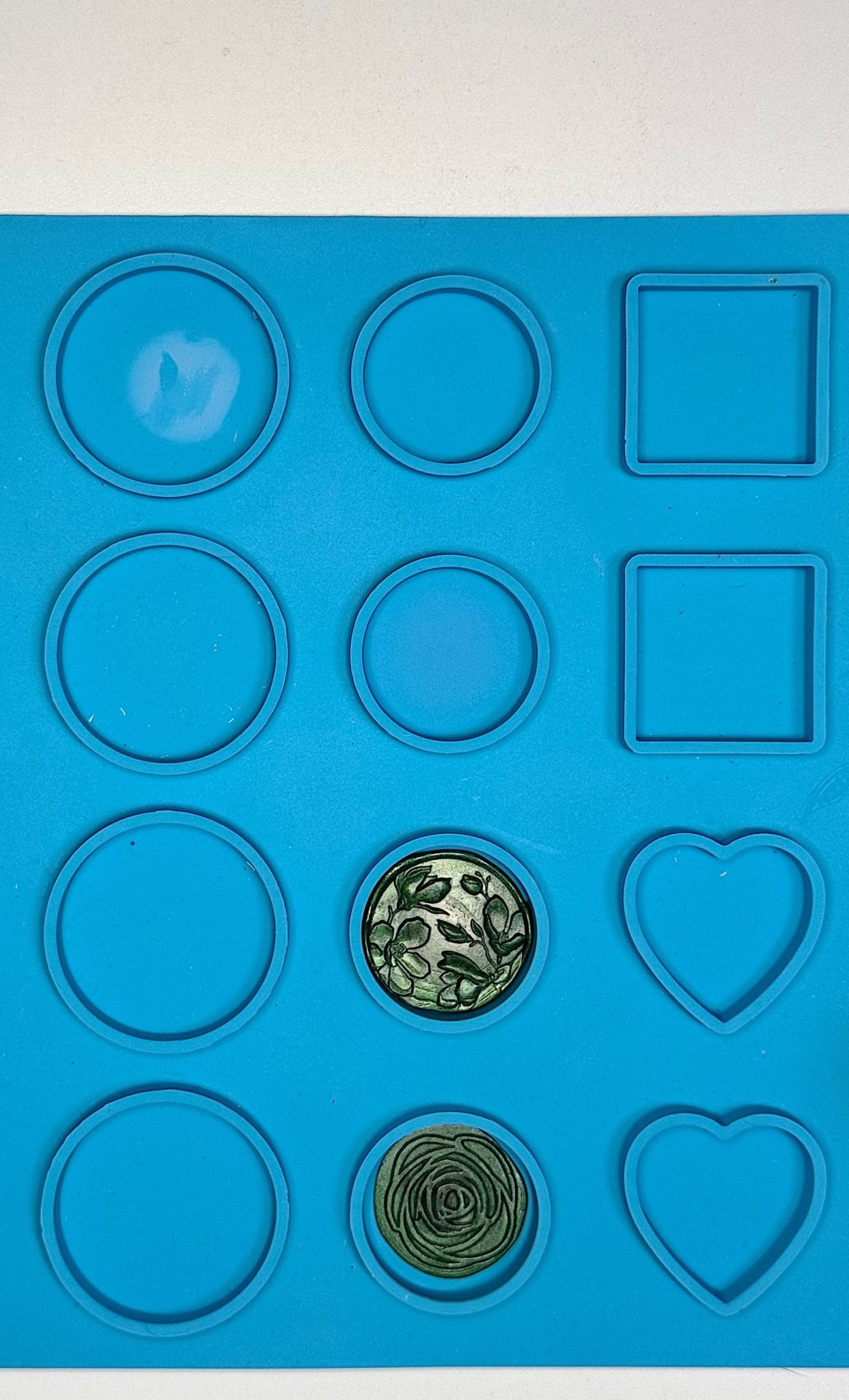 A blue silicone mold tray with circular, square, and heart-shaped cavities. Two of the circular cavities contain green designs, one with a floral pattern and one with a spiral shape.