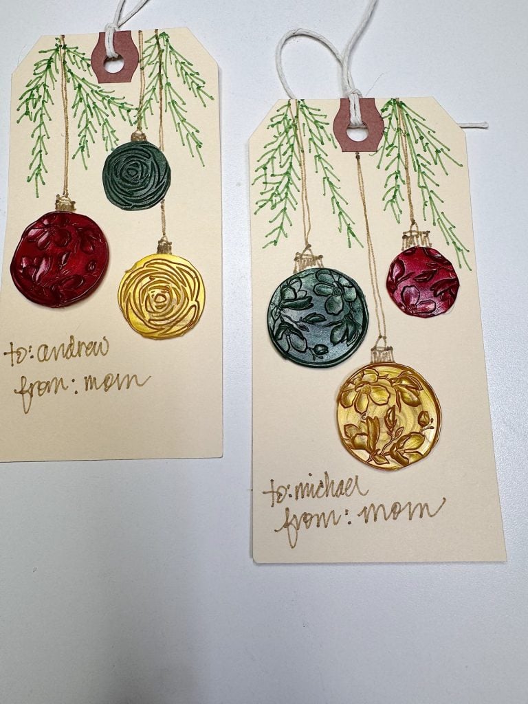 Two handmade gift tags decorated with embossed Christmas ornaments and green branches, labeled "to: andrew from: mom" and "to: michael from: mom" in cursive handwriting.