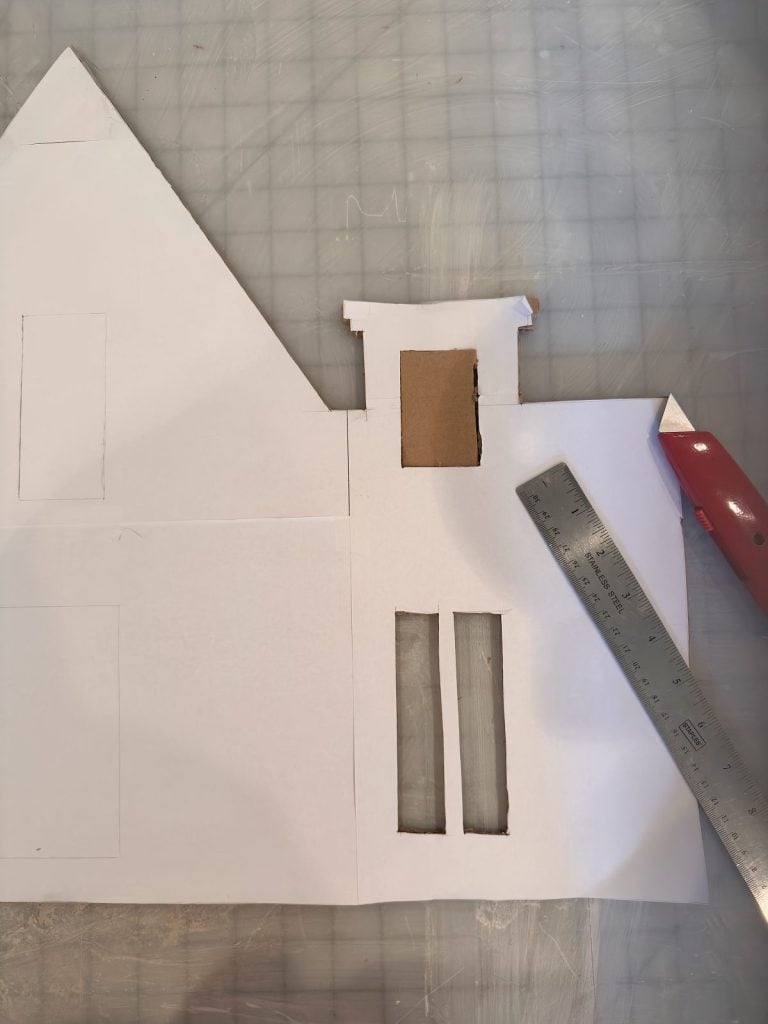 A piece of white paper with cutouts and drawn shapes lies on a grid surface, next to a metal ruler and a red utility knife.