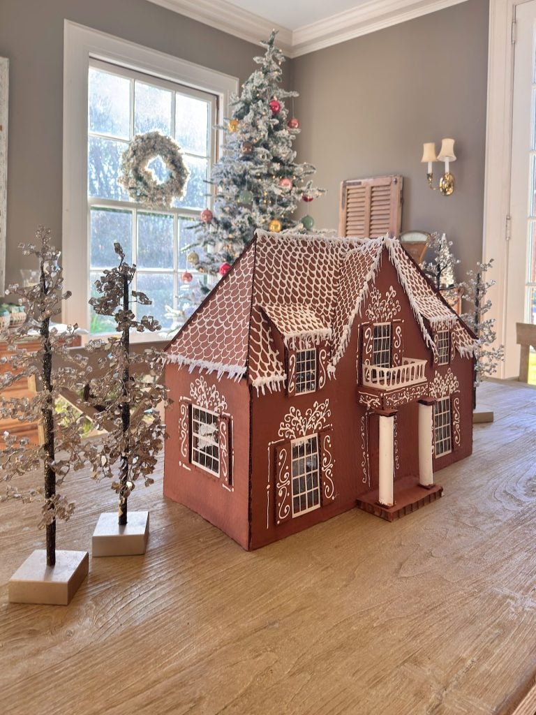 A gingerbread house decorated with white icing sits on a wooden table, with small artificial trees and a decorated Christmas tree in the background.