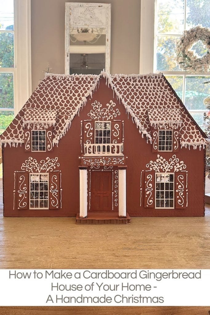 A cardboard gingerbread house model decorated with white icing designs is displayed indoors on a table.