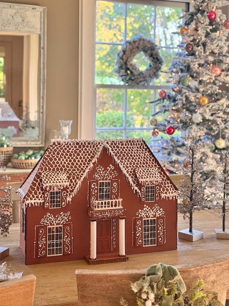 A detailed gingerbread house sits on a wooden table, with a decorated Christmas tree and a wreath visible in the background by a window.