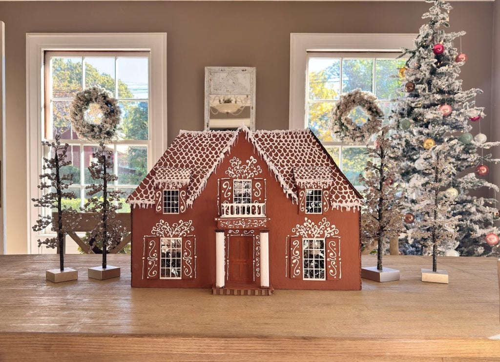 A large gingerbread house with white icing decorations sits on a wooden table, surrounded by small decorative trees and wreaths, with a Christmas tree in the background.
