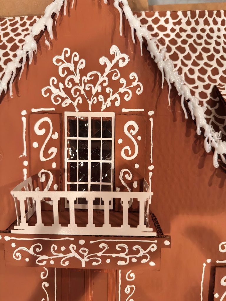 Close-up of a gingerbread house with white icing decorations, featuring a window and a small balcony, and icing resembling snow on the roof.