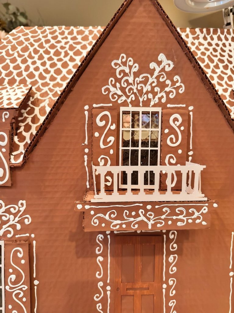 Close-up of a gingerbread house with white icing decorations, showing a balcony, window, and part of the roof with piped icing patterns.