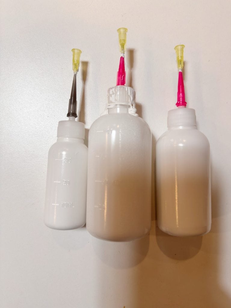 Three white plastic squeeze bottles with measurement markings, each fitted with a needle-tip cap, are arranged upright on a white surface.