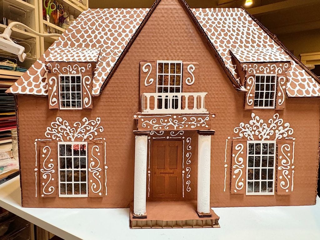 A large cardboard house model decorated to resemble a gingerbread house, with white icing-like patterns and detailed window and door designs.