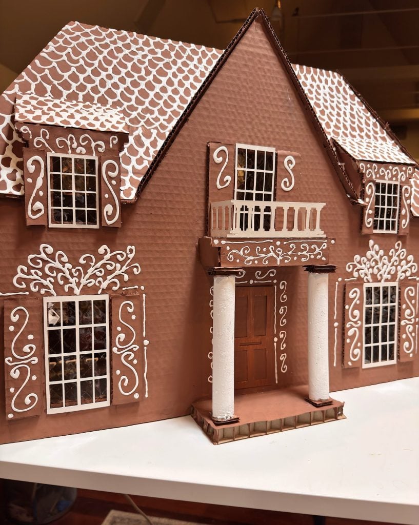 A large gingerbread house decorated with white icing patterns, featuring tall windows, a double front door, columns, and a balcony.