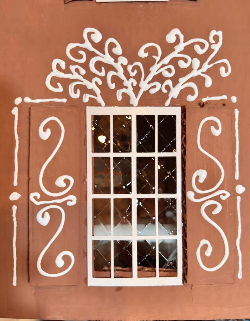 A rectangular window with white decorative scrollwork and accents on a brown wall, featuring a grid of glass panes and ornamental patterns above and beside it.