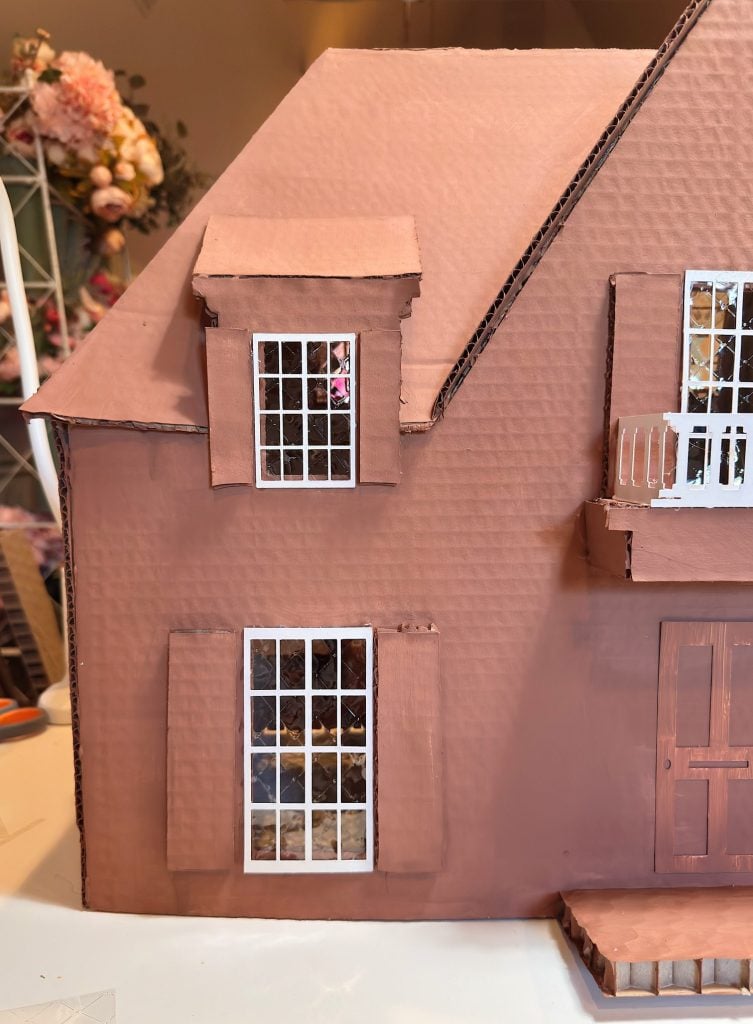 A close-up view of a cardboard model house featuring windows with white frames, a small balcony, and a brown painted exterior.