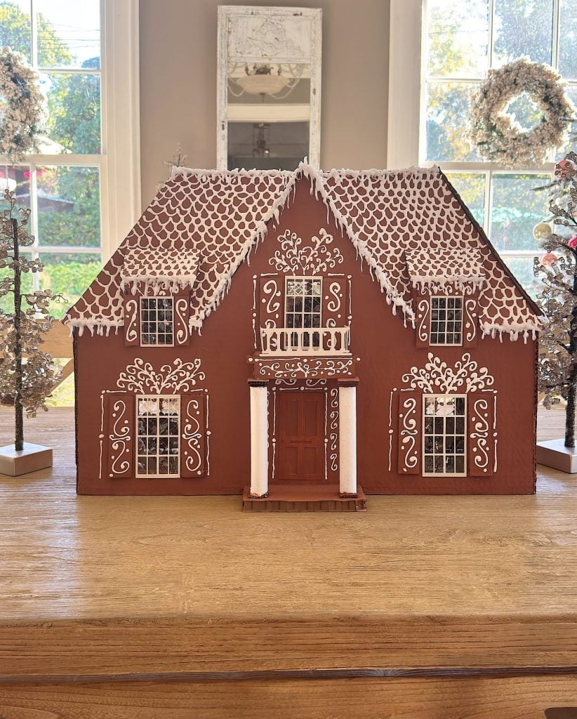 A gingerbread house decorated with white icing patterns sits on a wooden table, with wreaths and trees visible in the background through large windows.