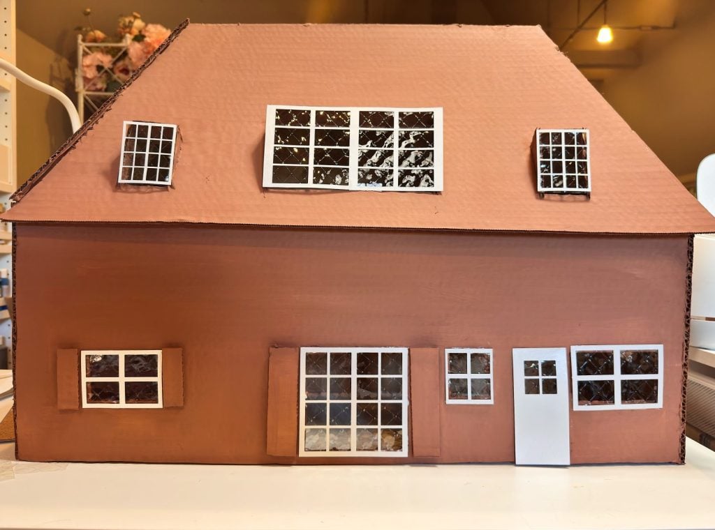 A cardboard model of a two-story brown house with white-framed windows and a white door, set indoors.