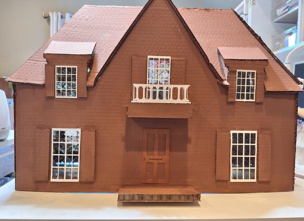 A handmade model of a brown two-story house with white-trimmed windows, a front door, a small balcony, and a slanted roof, displayed indoors.